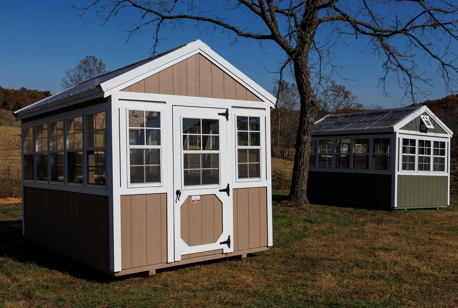 Amish-built greenhouse with glass panels and tan siding; a second greenhouse stands behind it